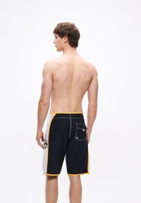 Černobílé kraťasy na koupání se žlutými akcenty, kontrastními bočními panely a zadní kapsou. Materiál vypadá lehký a vhodný na plavky.