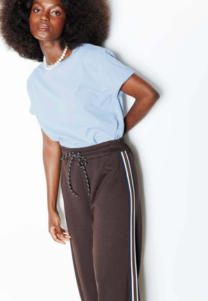 Femme aux cheveux afro portant un t-shirt bleu clair et un pantalon de survêtement marron avec des bandes latérales blanches et bleues, debout devant un fond blanc.