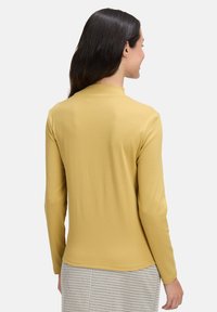 Żółta bluzka z długim rękawem, o wysoko stójce, gładkiej fakturze i dopasowanym kroju, zestawiona z spódnicą w kratę.