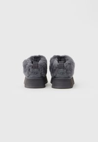 Grå UGG slip-on sko med tætsiddende pelsfor og sorte såler, set bagfra på en ensfarvet lys baggrund.