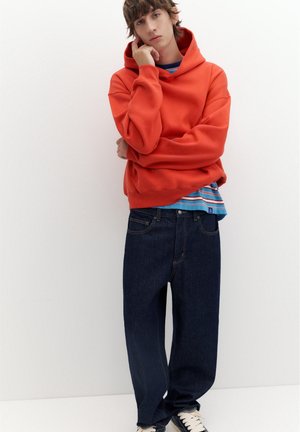 Jeune personne en sweat à capuche rouge, chemise rayée, jean large foncé et baskets, debout contre un mur blanc avec une main sur le visage.