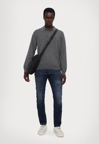 Graues langärmliges Polohemd, dunkelblaue Jeans und weiße Turnschuhe. Eine schwarze Umhängetasche wird über die Schulter getragen.