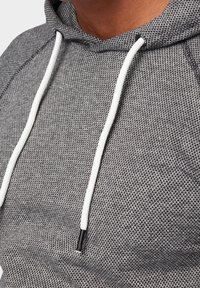 Grijze hoodie van een textuurstof, met witte trekkoorden en zwarte accenten. Close-up zicht laat het geweven patroon van het materiaal zien.