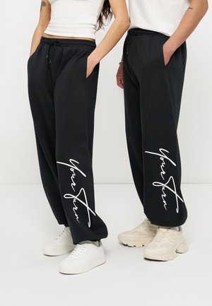 Dwie osoby w czarnych dresach z białym tekstem w stylu pisma na nogawkach, białe trampki, stoją z rękami w kieszeniach na białym tle.