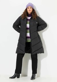 Svart puffjacka med dragkedja, med en huva och diagonala sömmar. Bärs över en flerfärgad randig tröja och svarta byxor.