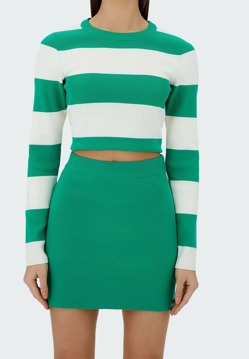 Femme portant un pull court à rayures horizontales vertes et blanches à manches longues, associé à une jupe verte unie taille haute.