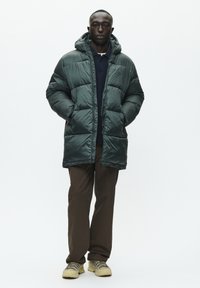 Homme portant une doudoune à capuche vert foncé, une chemise bleue marine, un pantalon marron et des chaussures de randonnée beiges, se tenant les mains dans les poches de sa veste.