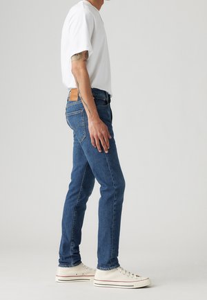 Uomo con sneakers bianche, jeans blu slim e una maglietta bianca semplice, in piedi di profilo su uno sfondo chiaro.