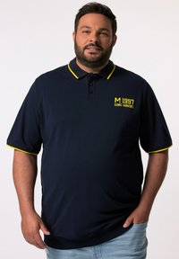 Mörkblå polo med gula detaljer. Har en tre-knapps placket och en tryckt logotyp på bröstet. Korta ärmar med kontrasterande kant.