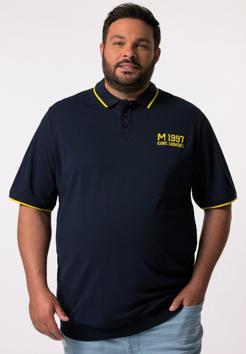 Mörkblå polo med gula detaljer. Har en tre-knapps placket och en tryckt logotyp på bröstet. Korta ärmar med kontrasterande kant.