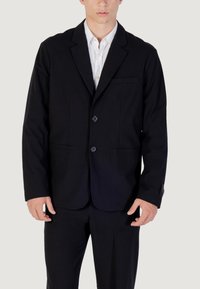 Uomo che indossa un blazer nero a due bottoni e pantaloni coordinati con una camicia bianca, in piedi contro uno sfondo chiaro e uniforme.