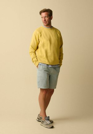 Man wearing yellow sweatshirt, light blue shorts, and gray sneakers standing with hands in pockets against beige background.