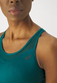 Mėlynas sportinis tanko tipo marškinėlis su tinklelio detalėmis, apvaliu kaklu ir logotipo akcentu ant krūtinės, rodantis glotnų audinį ir pritaikytą dizainą.
