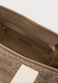 Beige kotte pehmea, tekstureeritud keha ja siledast nahast äär. Omab lukuga taskut ja kontrastset valget rihma.