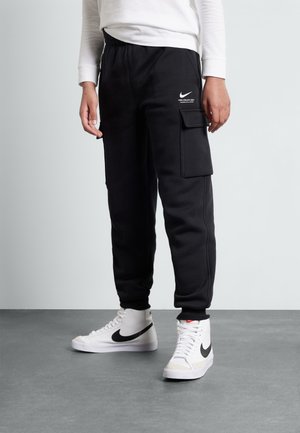 Personne portant un jogging cargo noir avec logo Nike et des baskets montantes blanches avec un swoosh noir Nike sur un sol gris.