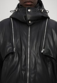 Veste en cuir noire avec un col haut, des zips argentés et plusieurs poches zippées sur la poitrine, avec une finition lisse et texturée.