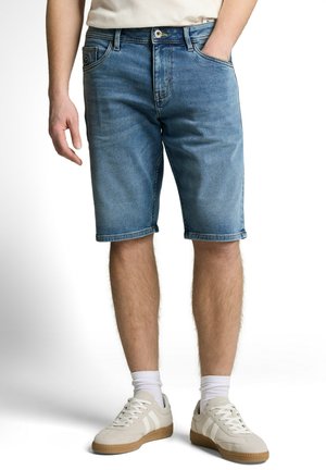 Mann trägt blaue Jeansshorts mittlerer Länge, weiße Socken und weiße Turnschuhe, steht mit einer Hand in der Hosentasche.