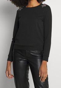 Svart sweatshirt med långa ärmar, rund halsringning och dekorativa detaljer på axlarna. Kombinerad med svarta läderbyxor. Slät textur.