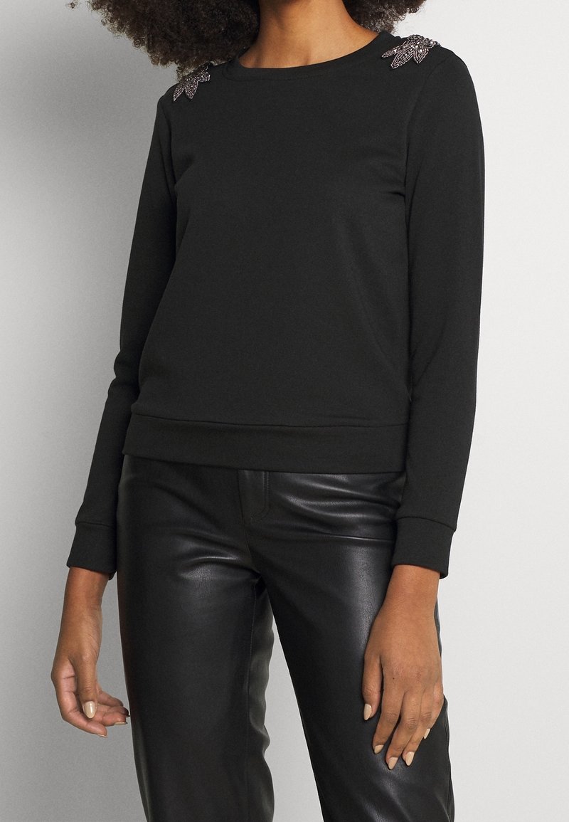 Svart sweatshirt med långa ärmar, rund halsringning och dekorativa detaljer på axlarna. Kombinerad med svarta läderbyxor. Slät textur.