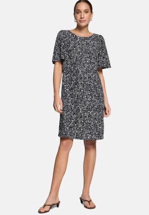 Kobieta w czarno-białej sukience do kolan z krótkimi rękawami i brązowych sandałach na niskim obcasie, stojąca z jedną nogą lekko wysuniętą do przodu.