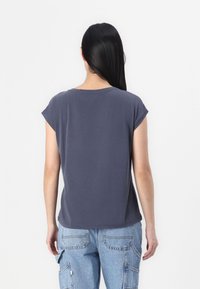 Žena s dlouhými černými vlasy, oblečená v tmavě šedém tričku s krátkým rukávem a světle modrých džínových cargo kalhotách, stojící zády.