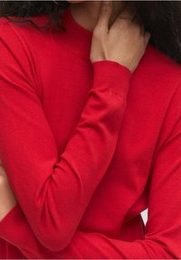 Haut rouge à manches longues en tissu doux avec un col rond. Il présente une coupe ajustée et une texture lisse, mettant en valeur la position des bras et des mains.