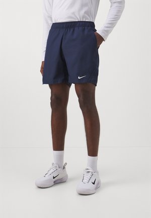 Modèle masculin portant un short Nike bleu marine, une chemise blanche à manches longues, des chaussettes blanches et des baskets blanches Nike, debout avec une main dans la poche.