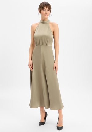 Frau steht in einem ärmellosen, olivgrünen Midikleid mit Neckholder und schwarzen, spitzen High Heels vor weißem Hintergrund.