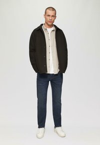 Schwarze Wolljacke über einem hellgrauen Hemd mit Knöpfen, weißem T-Shirt, dunkelblauen Jeans und weißen Sneakers. Schlichtes, modernes Design und Passform.