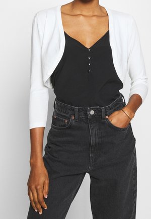 Vrouw met een witte cropped cardigan over een zwart topje met knopen, ingestopt in een zwarte high-waisted spijkerbroek, met één hand in de zak.