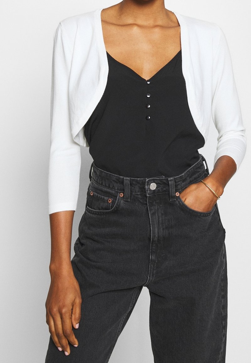 Femme portant un cardigan blanc court sur un top noir boutonné, rentré dans un jean noir taille haute, avec une main dans la poche.