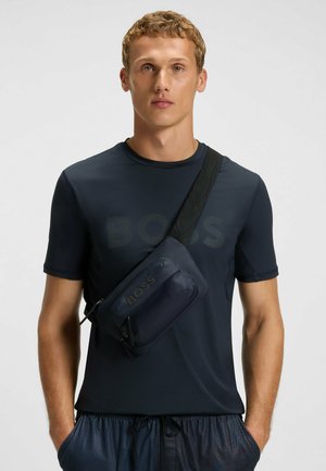 Navy crossbody bag with a smooth texture, zip closure, and embossed "BOSS" logo; worn over a navy short-sleeve athletic shirt.