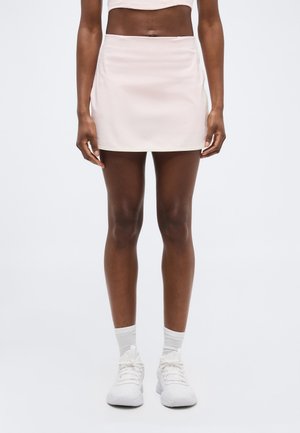 Γυναίκα που φοράει ανοιχτό ροζ αθλητική φούστα, λευκές κάλτσες και λευκά αθλητικά παπούτσια, όρθια μπροστά σε ένα απλό λευκό φόντο.