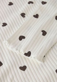 Valge tekstuuriga kangas, millel on vertikaalsed triibud ja väiksed tumedad sädeleved südame mustrid, mis on laiali üle pinna.