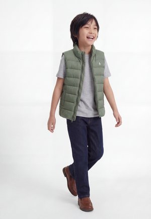 Mosolygós fiú sétál zöld steppelt mellényben, szürke pólóban, sötét farmerban és barna cipőben, fehér háttér előtt.