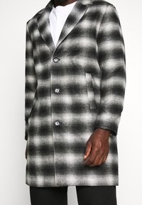 Cappotto a scacchi bianco e nero, realizzato in tessuto testurizzato, con colletto a pistagna, sei bottoni e tasche laterali.