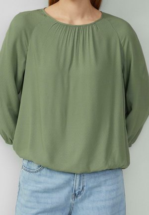 Personne portant un haut ample vert olive à manches longues avec un col froncé et un jean en denim bleu clair, debout devant un fond uni.