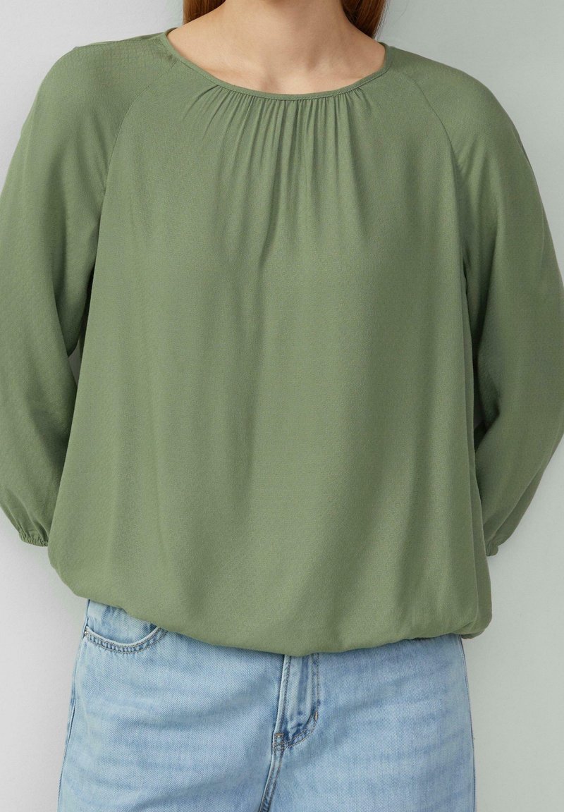 Personne portant un haut ample vert olive à manches longues avec un col froncé et un jean en denim bleu clair, debout devant un fond uni.