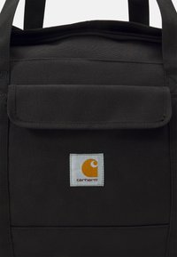 Čierna taška cez rameno vyrobená z odolnej tkaniny, ktorá má klopový uzáver, horné uchá a bielu štvorcovú nášivku s oranžovým logom.