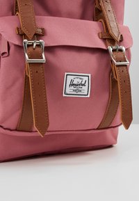 Sac à dos rose avec des accents en cuir marron, présentant des bretelles doublées de boucles en argent et un patch logo Herschel blanc. Tissu texturé.