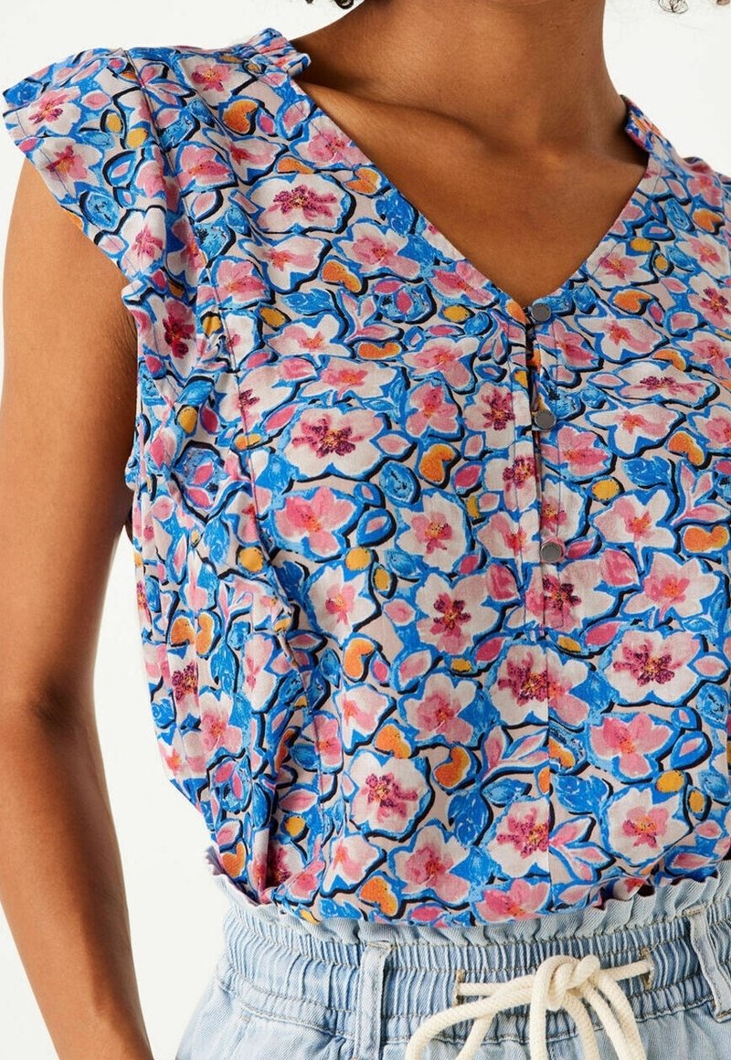 Blouse fleurie en rose, bleu et jaune avec manches à volants, encolure en V, boutons et motif complexe sur un tissu léger.