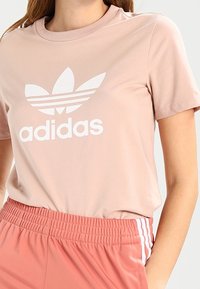 Ljusrosa bomulls t-shirt med korta ärmar, rund hals och stor vit Adidas-logotyp med tre ränder och bladdesign.