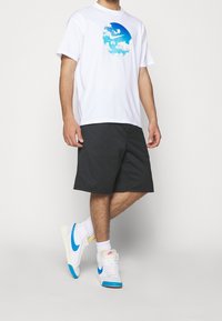 Bílá bavlněná trička s modro-bílou grafikou mraků, spárované s černými šortkami a bílými teniskami s modrými akcenty a žlutými detaily.