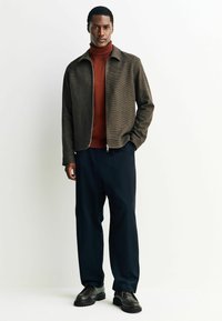 Braune, strukturierte Zip-Jacke über einem roten Rollkragenpullover, kombiniert mit lockeren schwarzen Hosen und schwarzen Schnürschuhen, vor einem weißen Hintergrund.