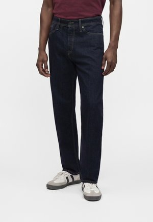 Homme portant un jean droit bleu foncé, des baskets blanches avec des rayures noires et un t-shirt rouge foncé, debout devant un fond clair uni.