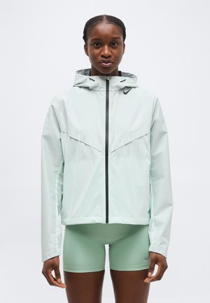 Naine, kes kannab helerohelist lukuga kapuutsiga jopet ja sobivaid lähedaltistuvaid lühikesi pükse, seisab ühevärvilise valge tausta ees.