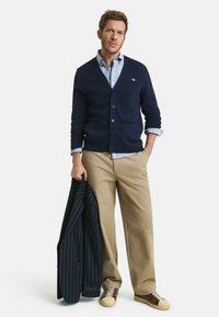 Marinefarbener Cardigan mit Knöpfen und Taschen, getragen über einem hellblauen Hemd. Beige Hosen und braune und cremefarbene Sneaker. Mann hält ein gestreiftes Sakko.