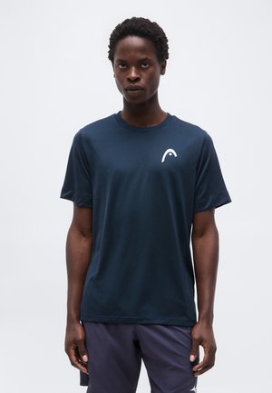 Mies seisoo päällään tummansininen lyhythihainen urheilupaita, jossa on valkoinen logo rinnassa, ja tummat urheilushortsit, yksiväristä taustaa vasten.