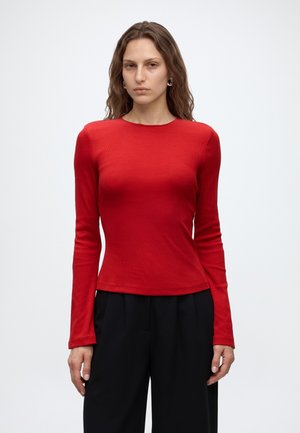 Femme aux longs cheveux bruns, portant un haut rouge ajusté à manches longues et un pantalon noir ample, debout devant un fond blanc uni.