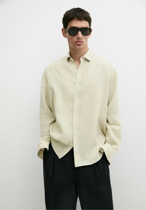 Ung mand iført sorte solbriller, løs beige skjorte med knapper og sorte bukser, stående med hænderne i lommerne mod en ensfarvet hvid baggrund.
